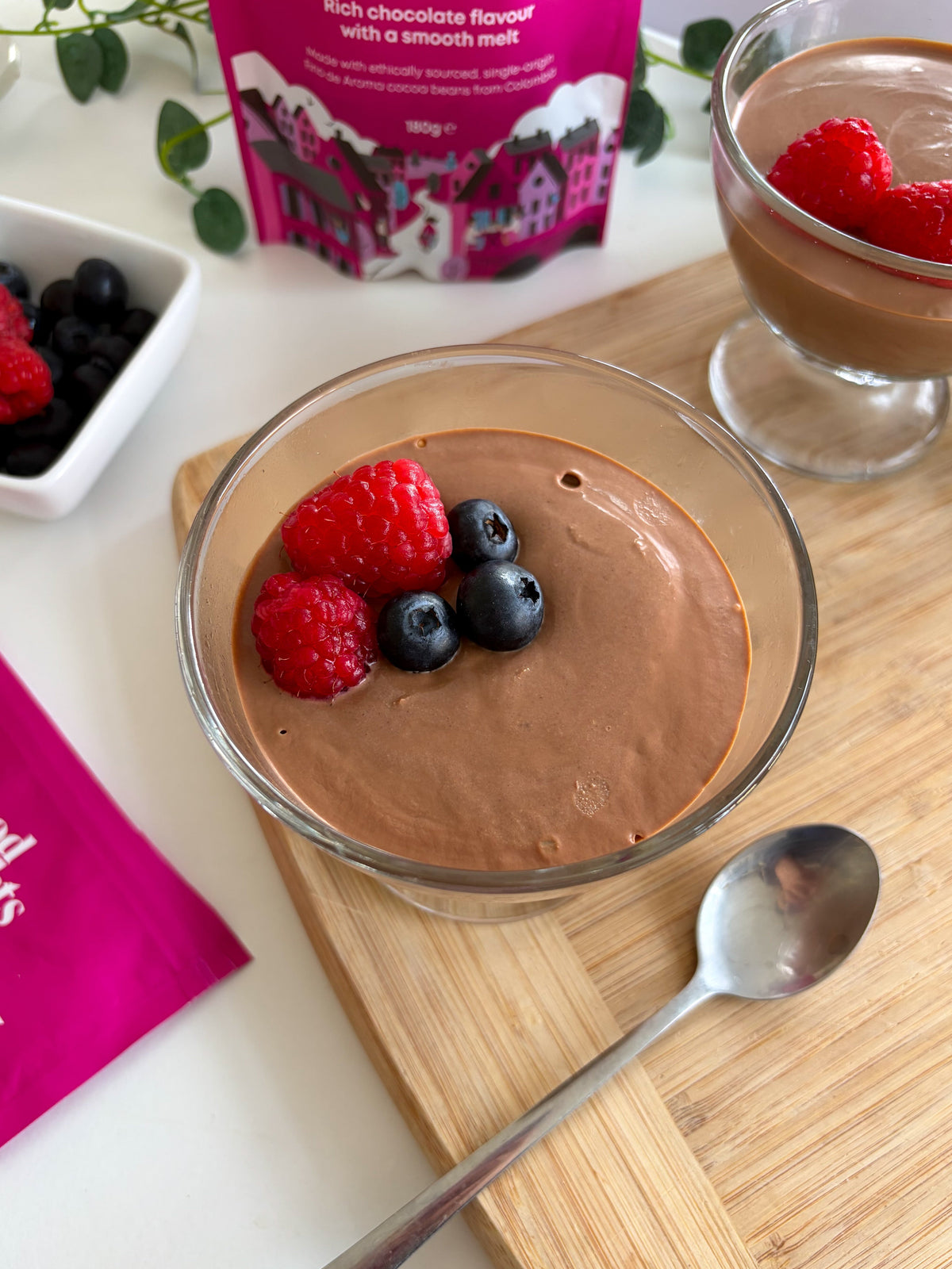 A glass of chocolate tofu mousse topped with raspberries and blueberries, next to a bag of Food Thoughts 70% Dark Chocolate Chips.