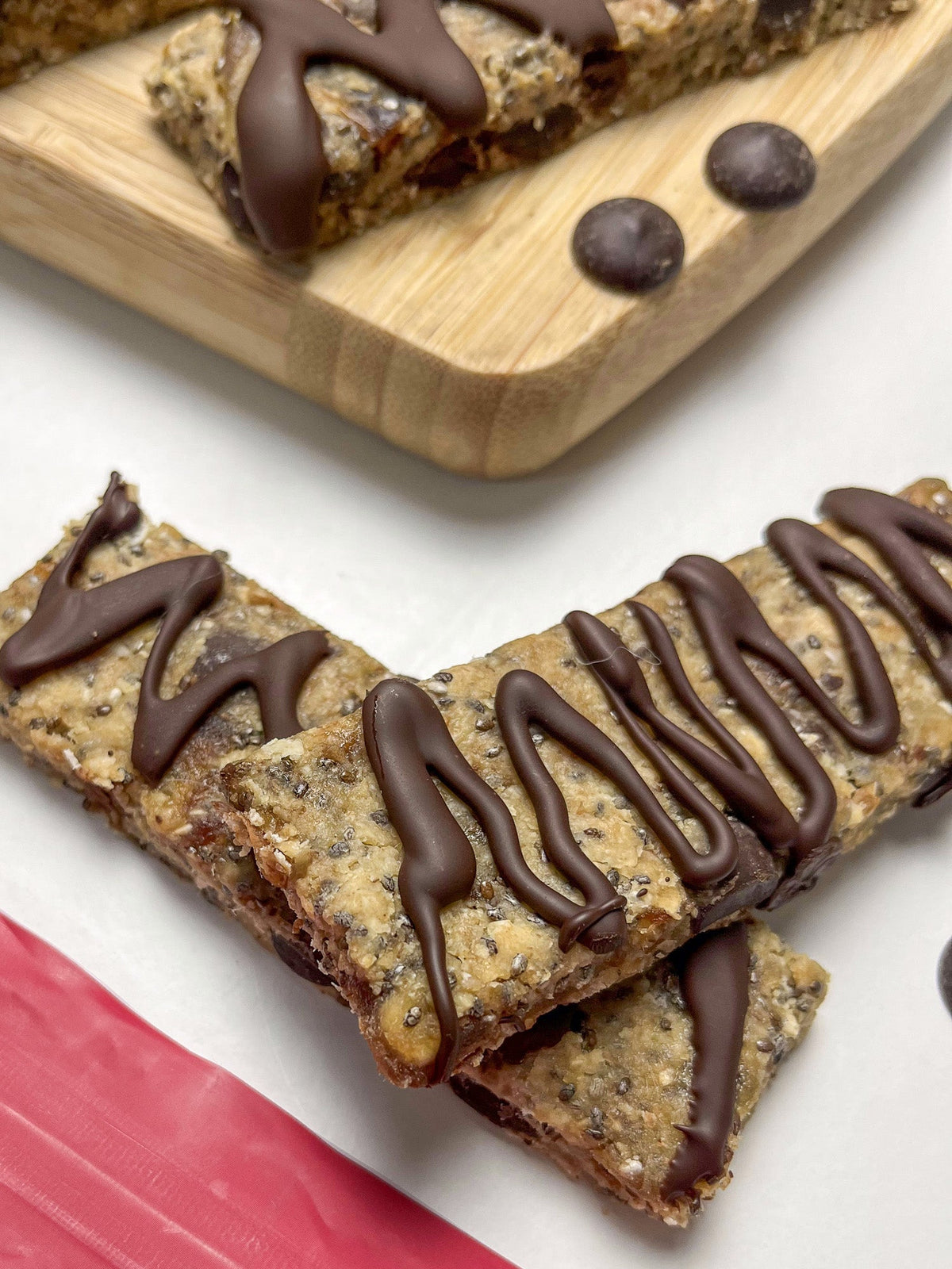 A row of chocolate-drizzled date granola bars on a wooden board next to a bag of Food Thoughts 70% Dark Chocolate Chips.